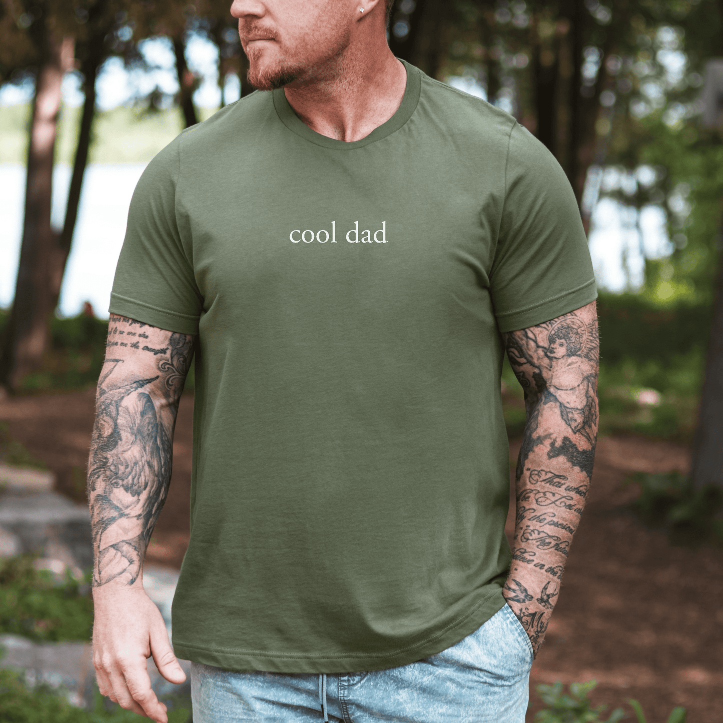 Tattooed man wearing olive green Minimalist Cool Dad Shirt with white centered cool dad text outdoors by lake