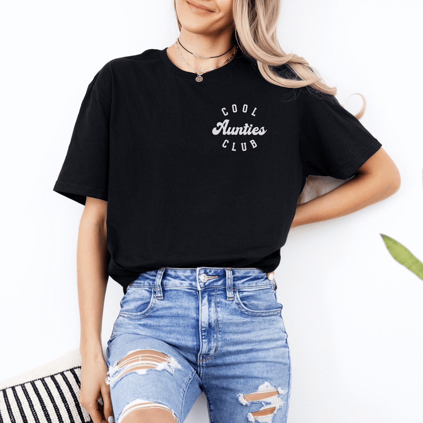 Woman wearing black tee with white Cool Aunties Club Shirt . logo paired with ripped light blue jeans and necklaces