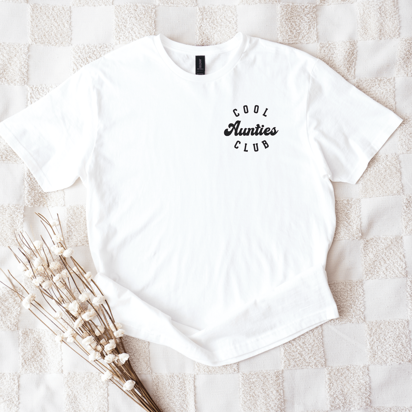 White cotton tee with small black Cool Aunties Club Shirt . chest logo styled on neutral textured blanket