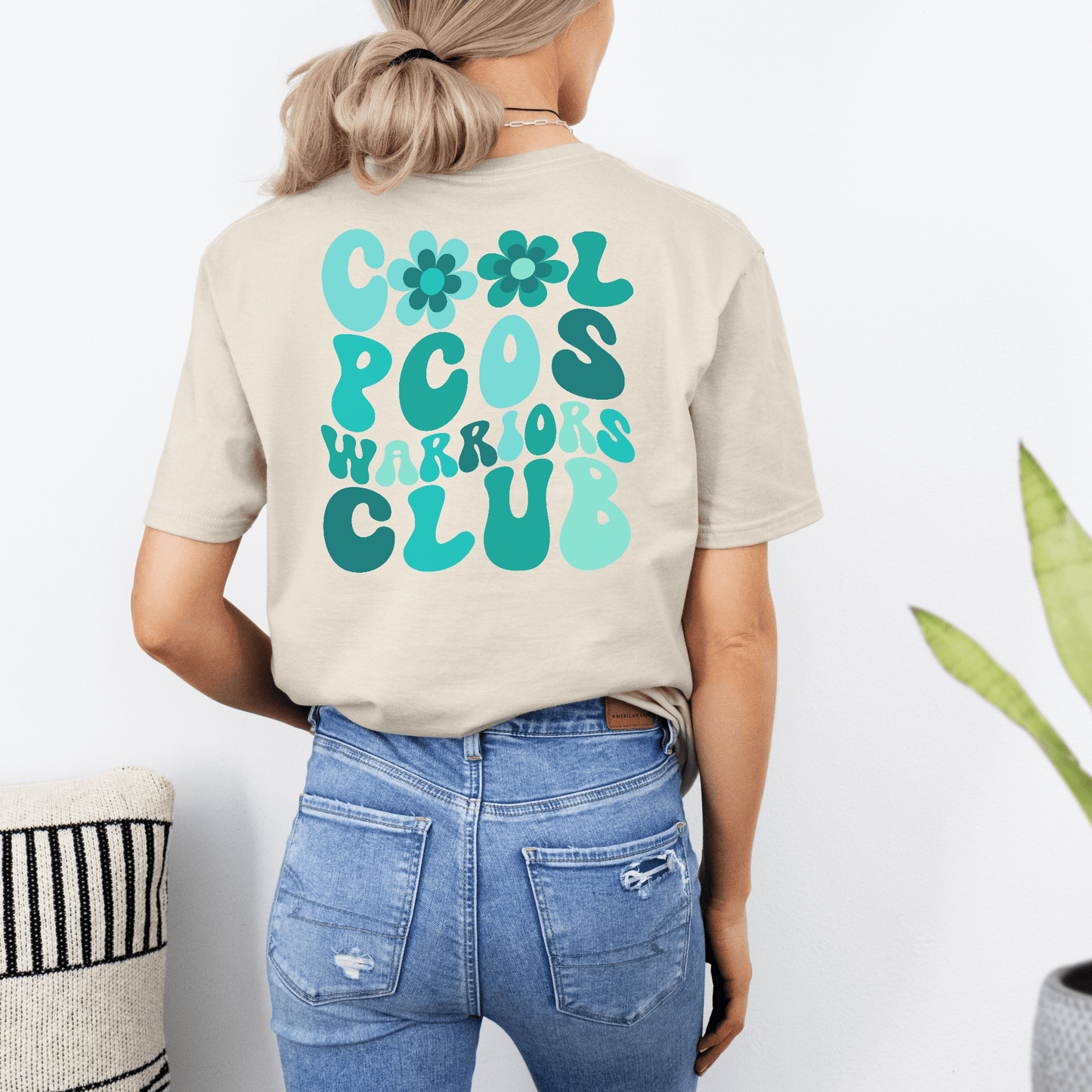 Woman wearing beige PCOS Awareness Shirt with teal retro floral Cool PCOS Warriors Club design on back and denim jeans