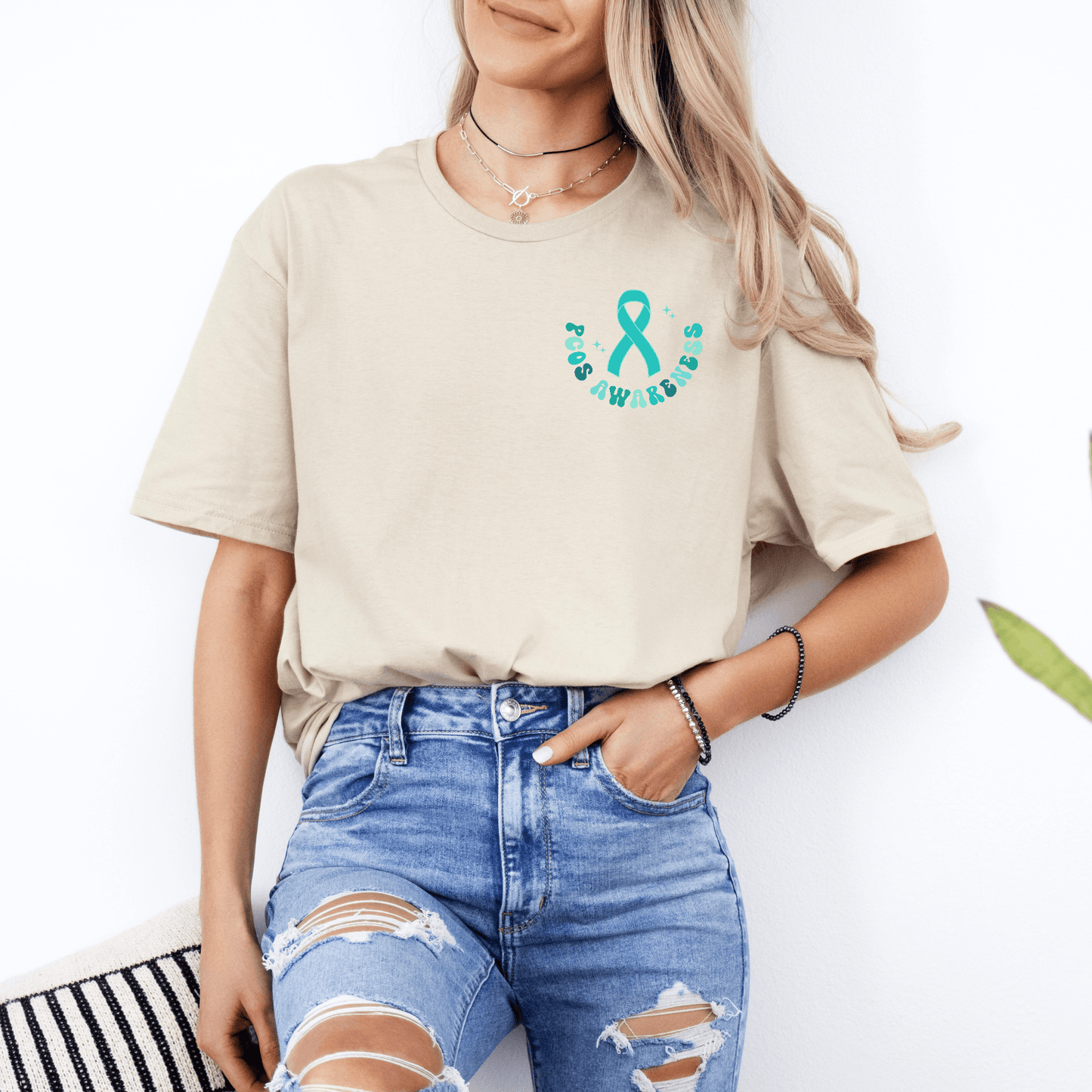 Woman wearing beige PCOS Awareness Shirt with teal ribbon chest logo tucked into ripped blue jeans casual style