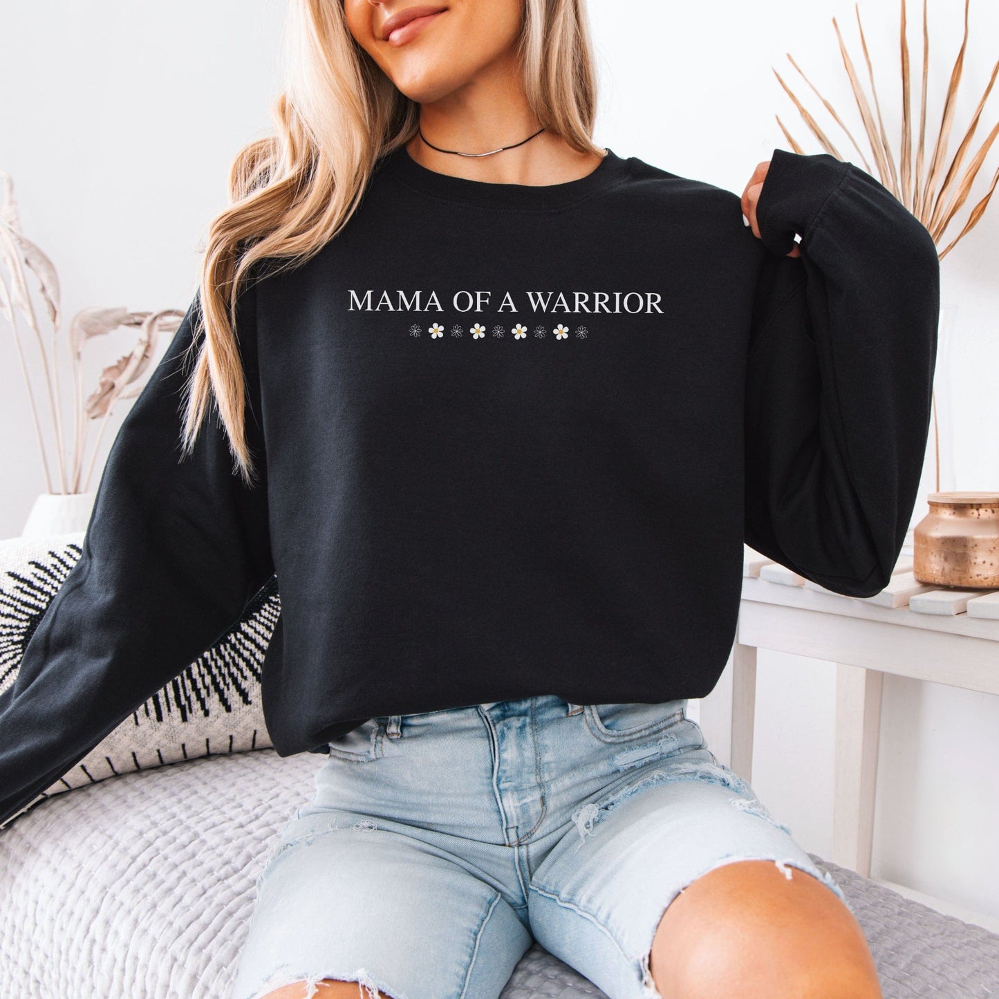 Woman wearing black Mama of a Warrior Sweatshirt with floral accents paired with distressed light denim jeans