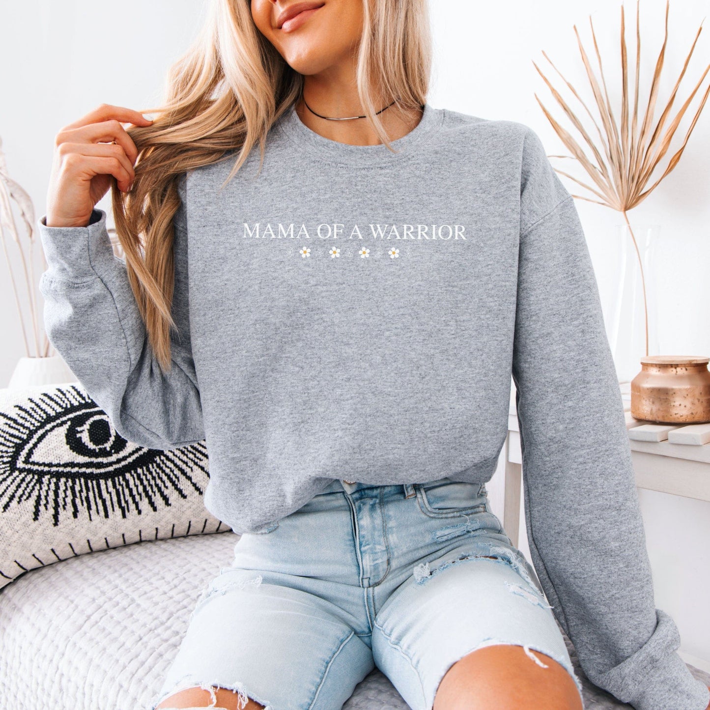 Woman wearing gray Mama of a Warrior Sweatshirt by Maddy and George Designs seated on couch with jeans