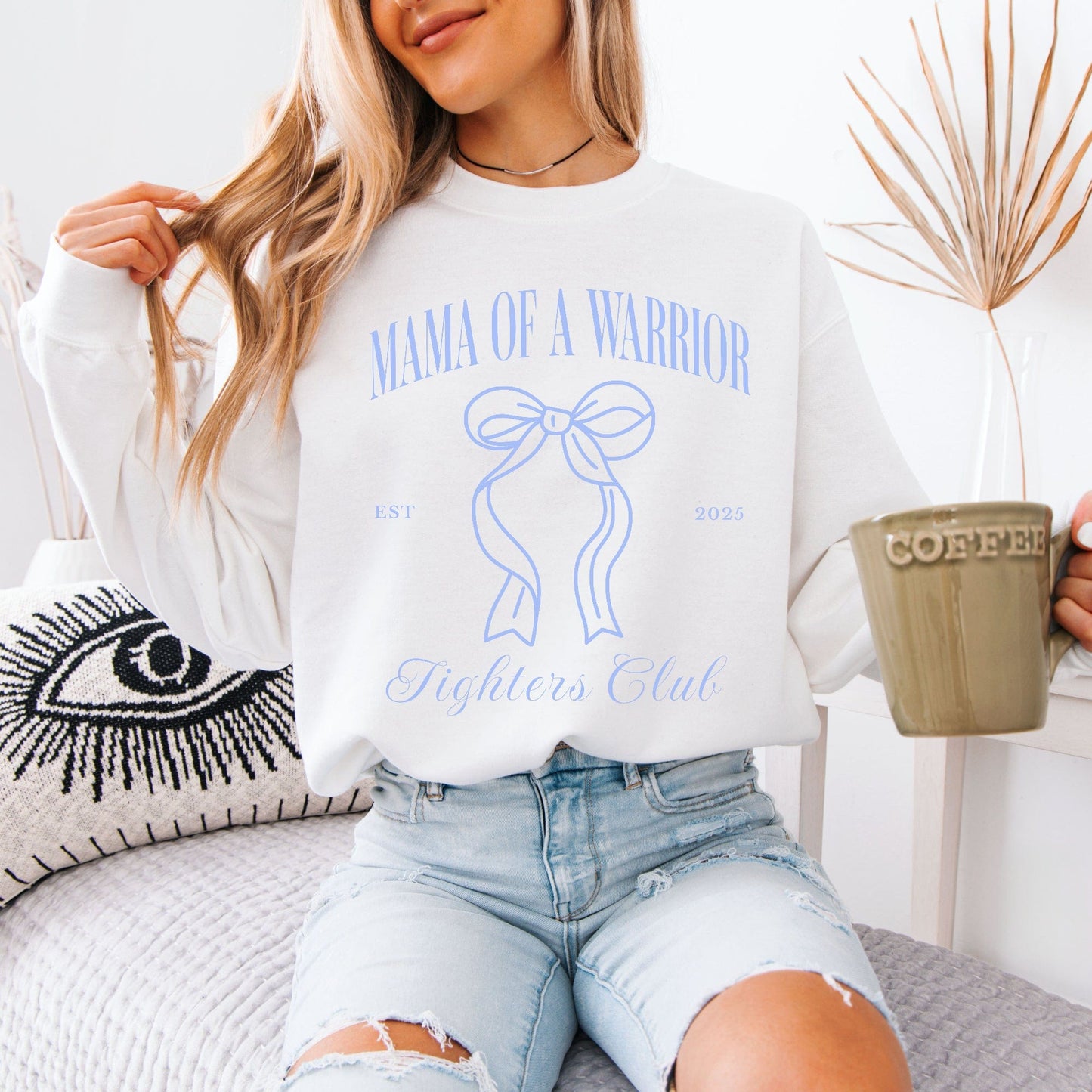 White sweatshirt with blue Mama of a Warrior print worn by woman holding coffee Medical Mom Gift