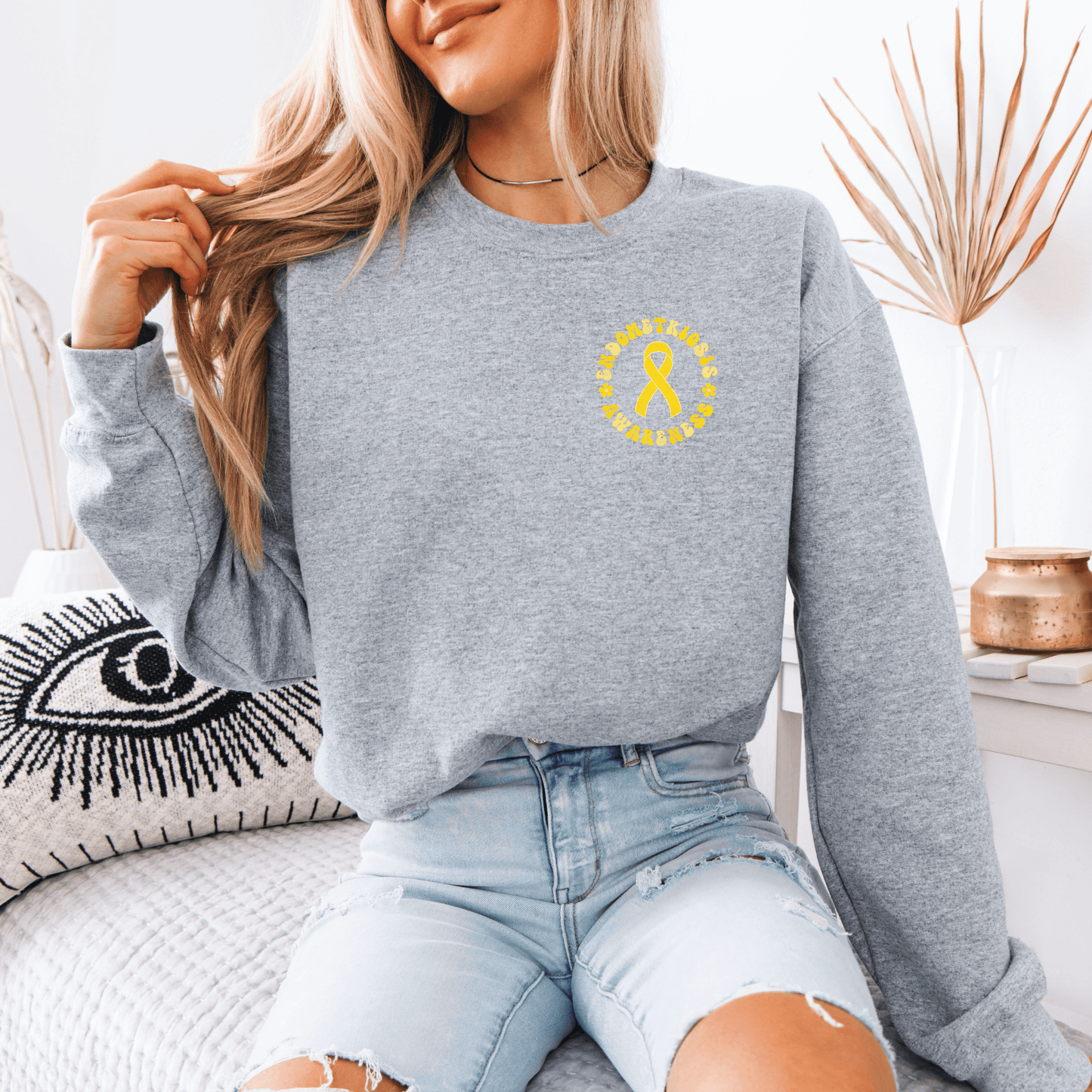 Young woman wearing gray Endometriosis Awareness Sweatshirt with small yellow ribbon on chest sitting on couch