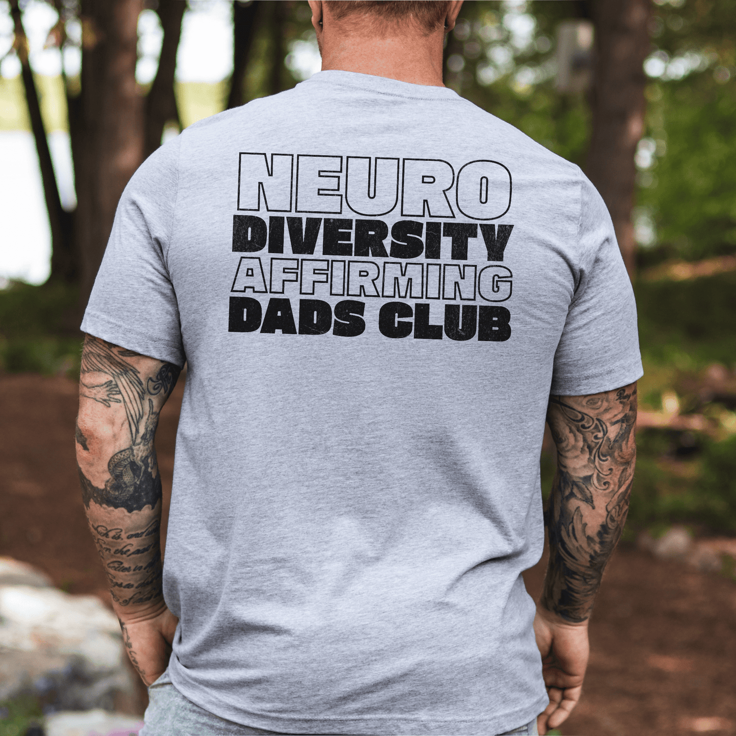 Back view of gray tee with bold black print reading Neurodiversity Affirming Dads Club Shirt worn by a tattooed dad outdoors