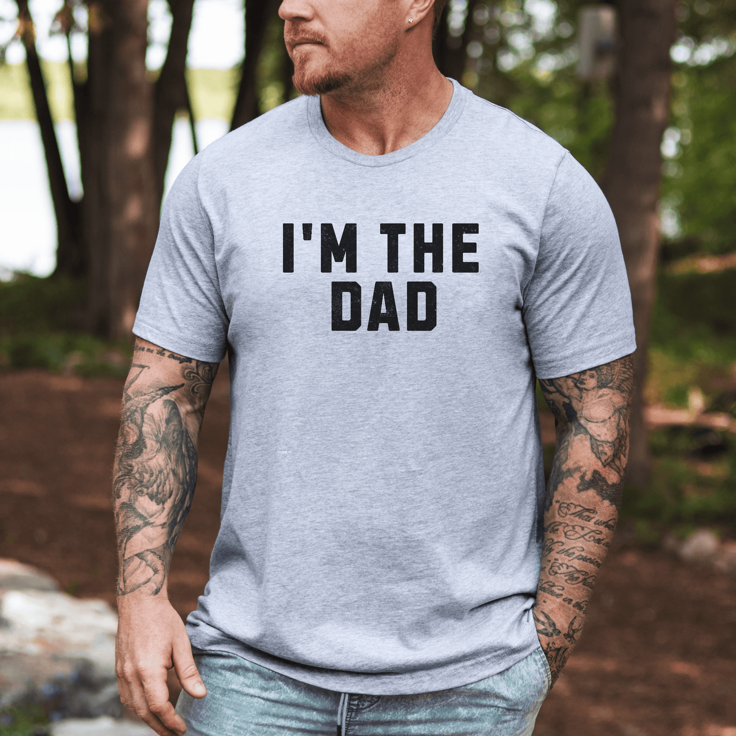 Man with tattooed arms wearing light gray I'm The Dad Shirt outdoors near trees and lake