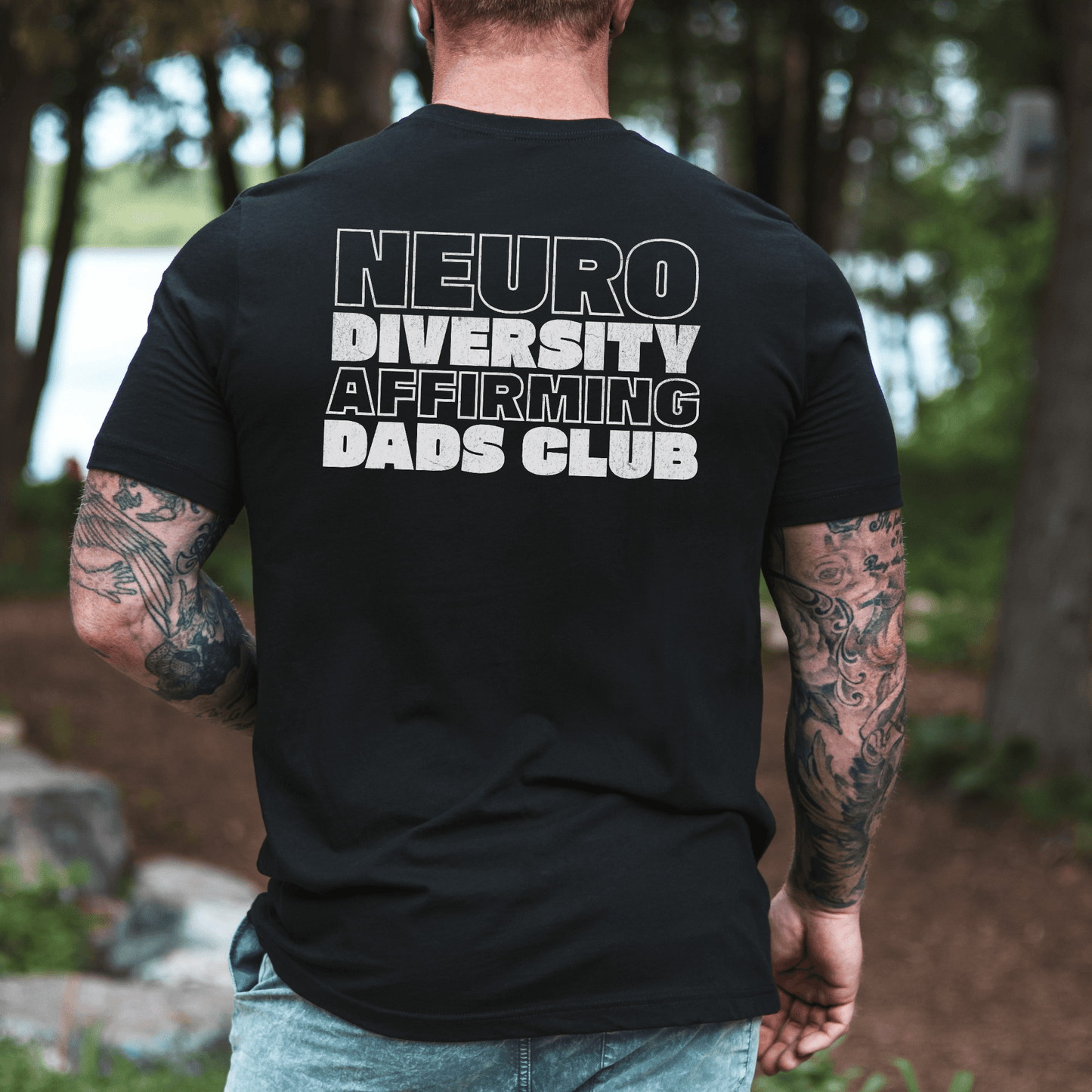 Back view of tattooed dad wearing black Neurodiversity Affirming Dads Club Shirt . bold white lettering outdoor setting