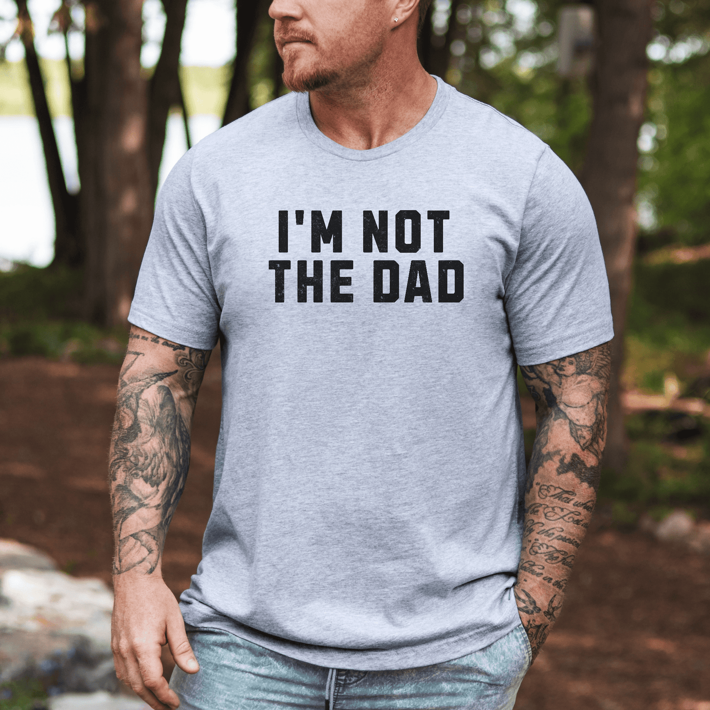 Tattooed man wearing a light gray funny I'm not the dad shirt with bold black text outdoors