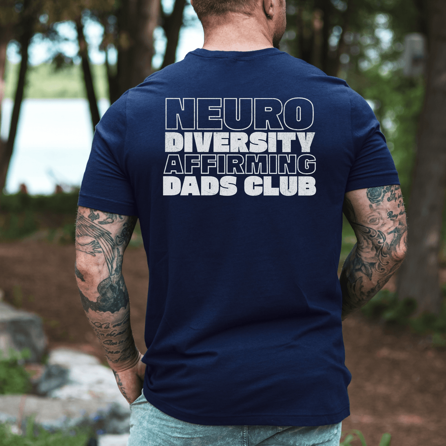 Man with tattooed arms wearing navy Neurodiversity Affirming Dads Club Shirt . back view outdoors by lake