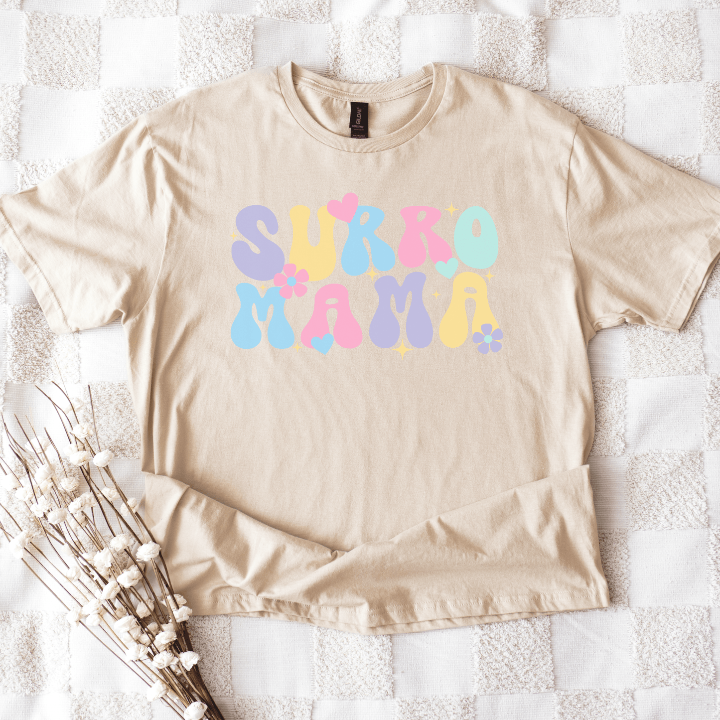 Cute Surrogate Mama Shirt beige tee with pastel retro bubble lettering hearts and flowers styled flat on cozy white quilt