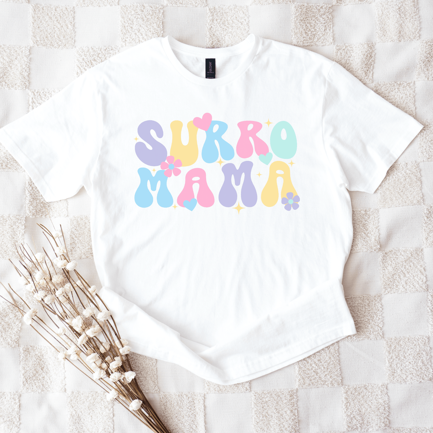 White t shirt with pastel bubble letters and flowers reading Surro Mama laid flat on blanket Cute Surrogate Mama Shirt