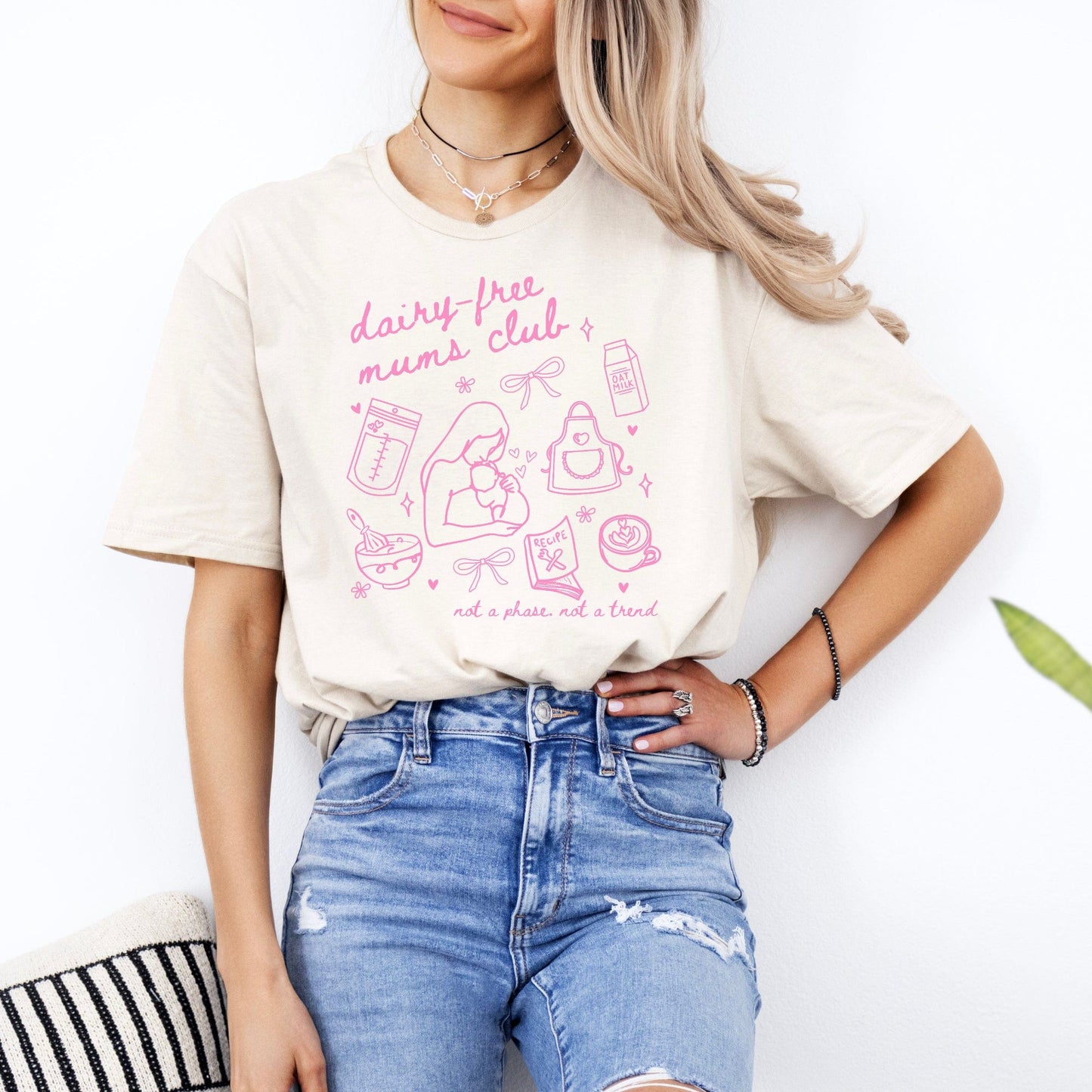 Woman wearing cream tee with pink graphic reading Dairy Free Mums Club Shirt showing breastfeeding icons and bottles