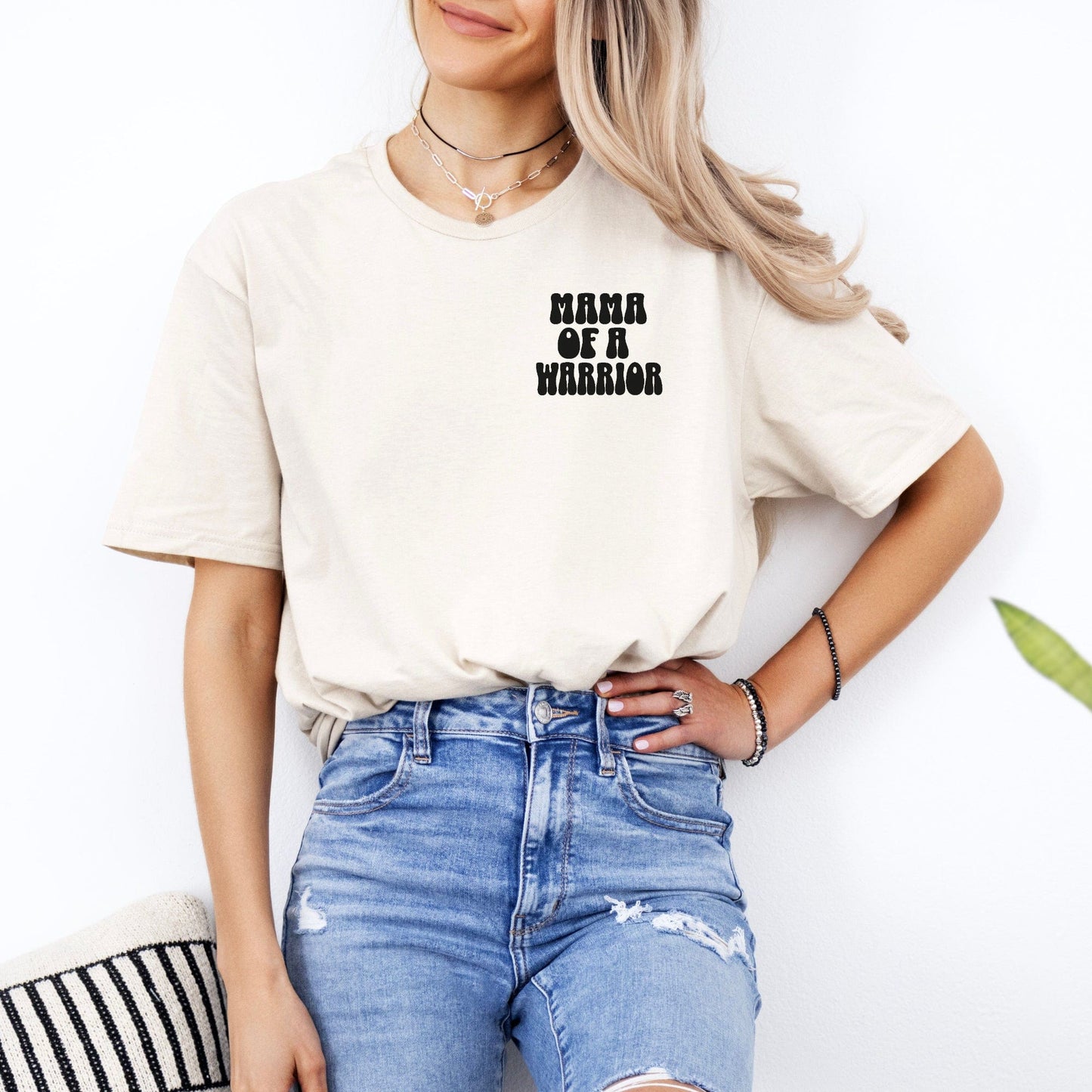 Mama of a Warrior Club Shirt cream tee worn by woman with layered necklaces and distressed jeans