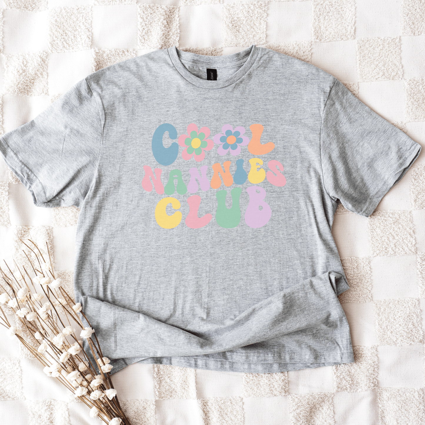 Gray unisex t shirt laid flat on blanket with pastel retro bubble lettering and flower graphics Cool Nannies Club Shirt
