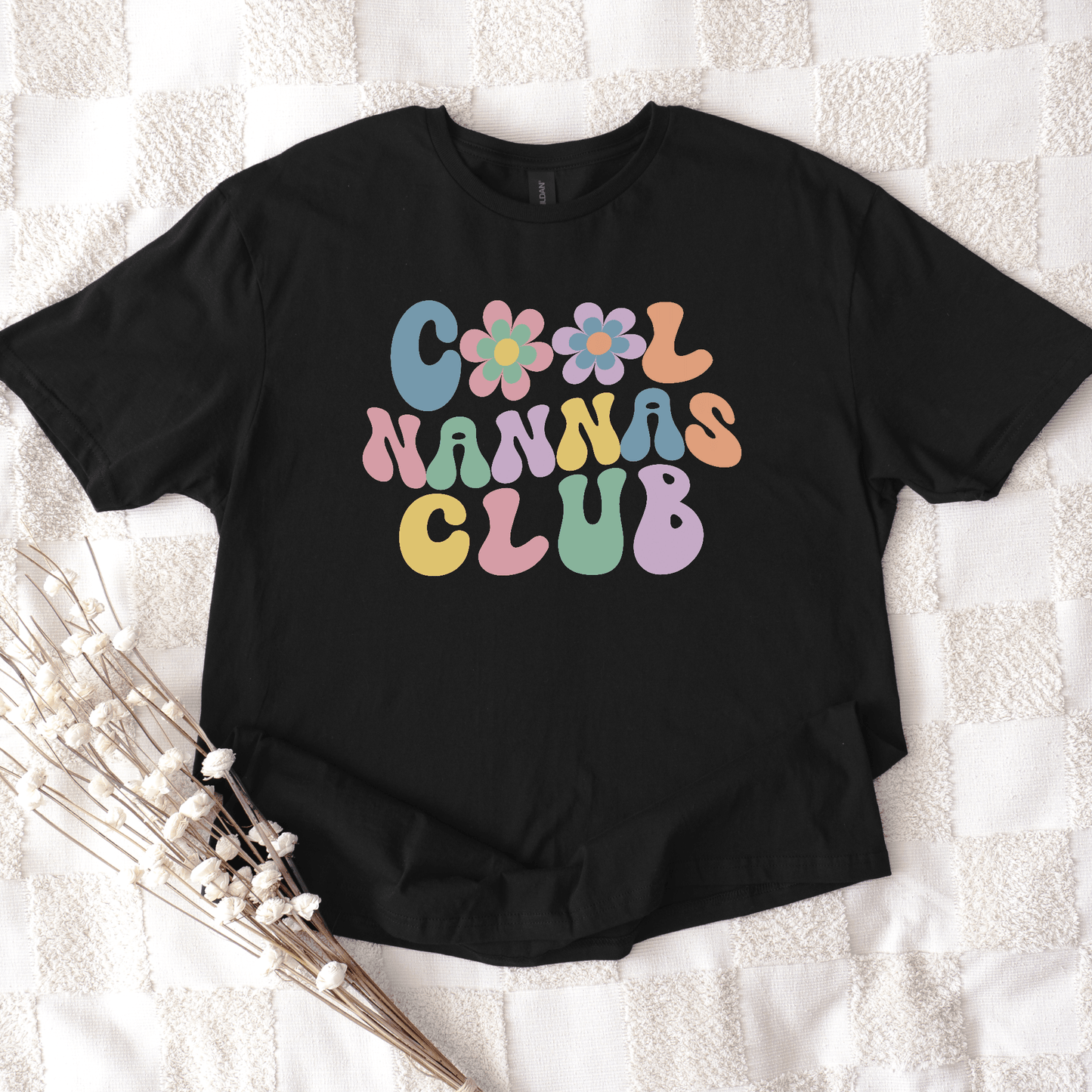 Black t shirt with retro pastel bubble letters and flowers reading Cool Nannas Club Shirt flat on white textured background