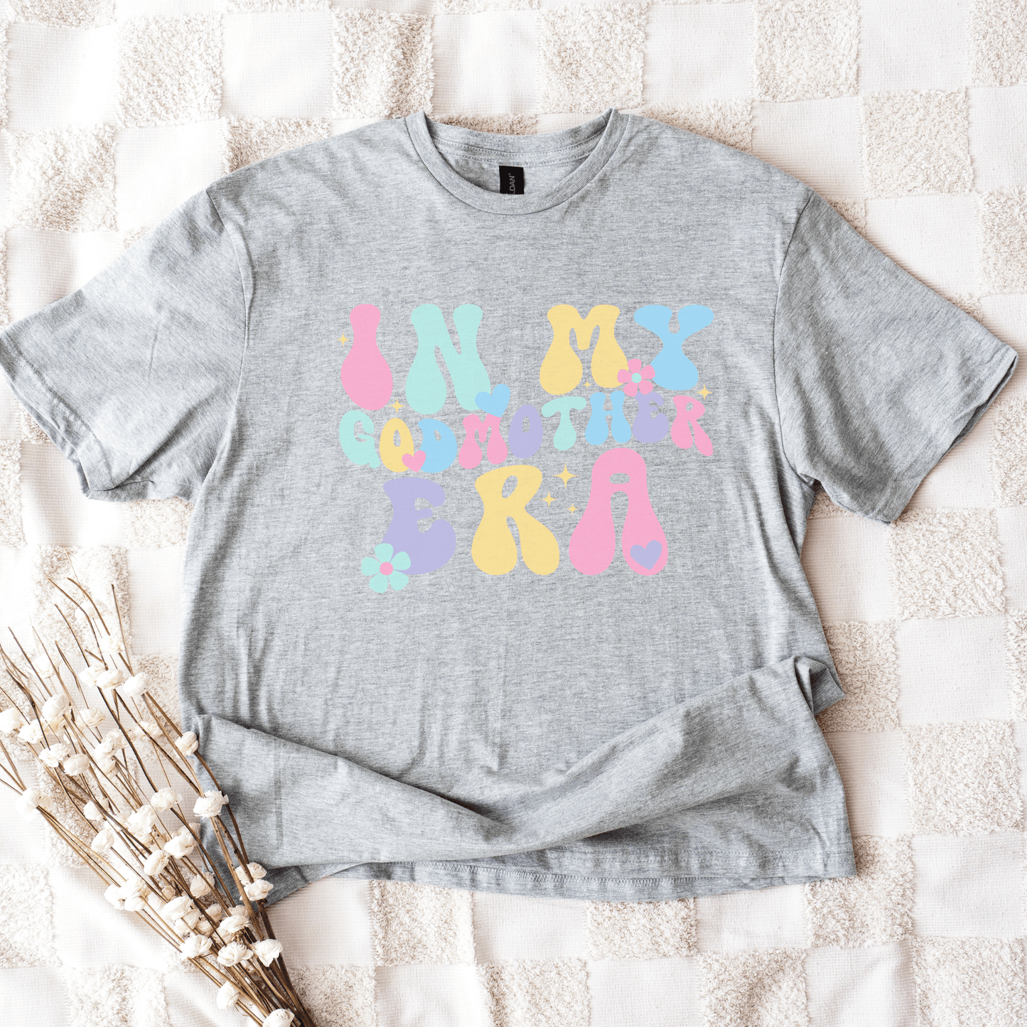 Gray unisex tee laid flat with pastel bubble letters and floral accents reading In My Godmother Era Godmother Announcement Shirt