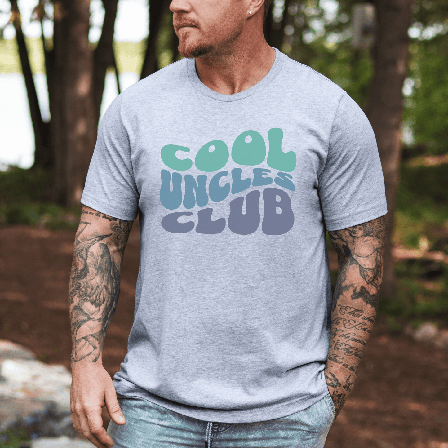 Man wearing light gray Cool Uncles Club T-Shirt . with retro green and blue bubble text outdoors