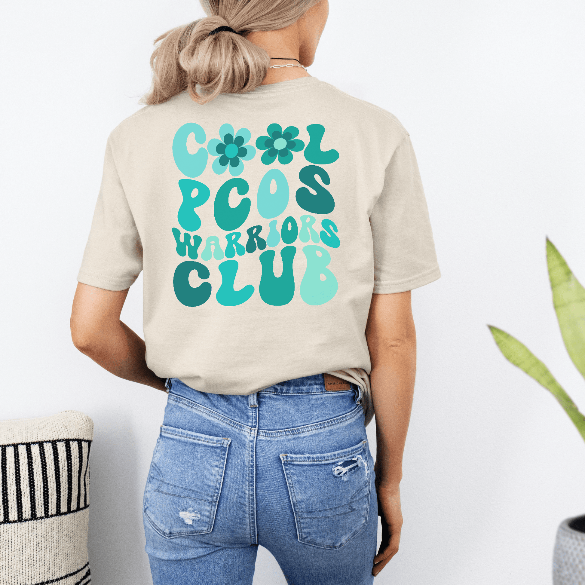 Woman wearing beige PCOS Awareness Shirt with teal retro floral Cool PCOS Warriors Club design on back and denim jeans
