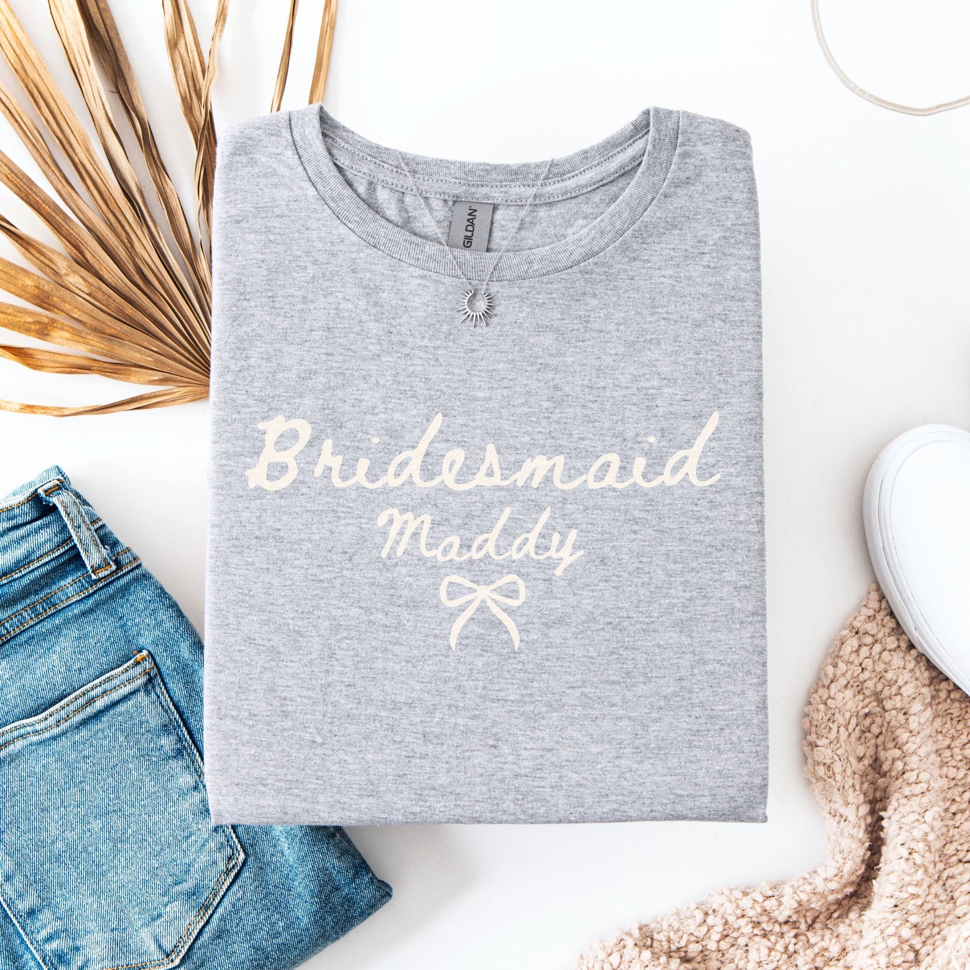 Gray folded Custom Bridesmaid T-Shirt printed with cream Bridesmaid Maddy script, styled with jeans and sneakers