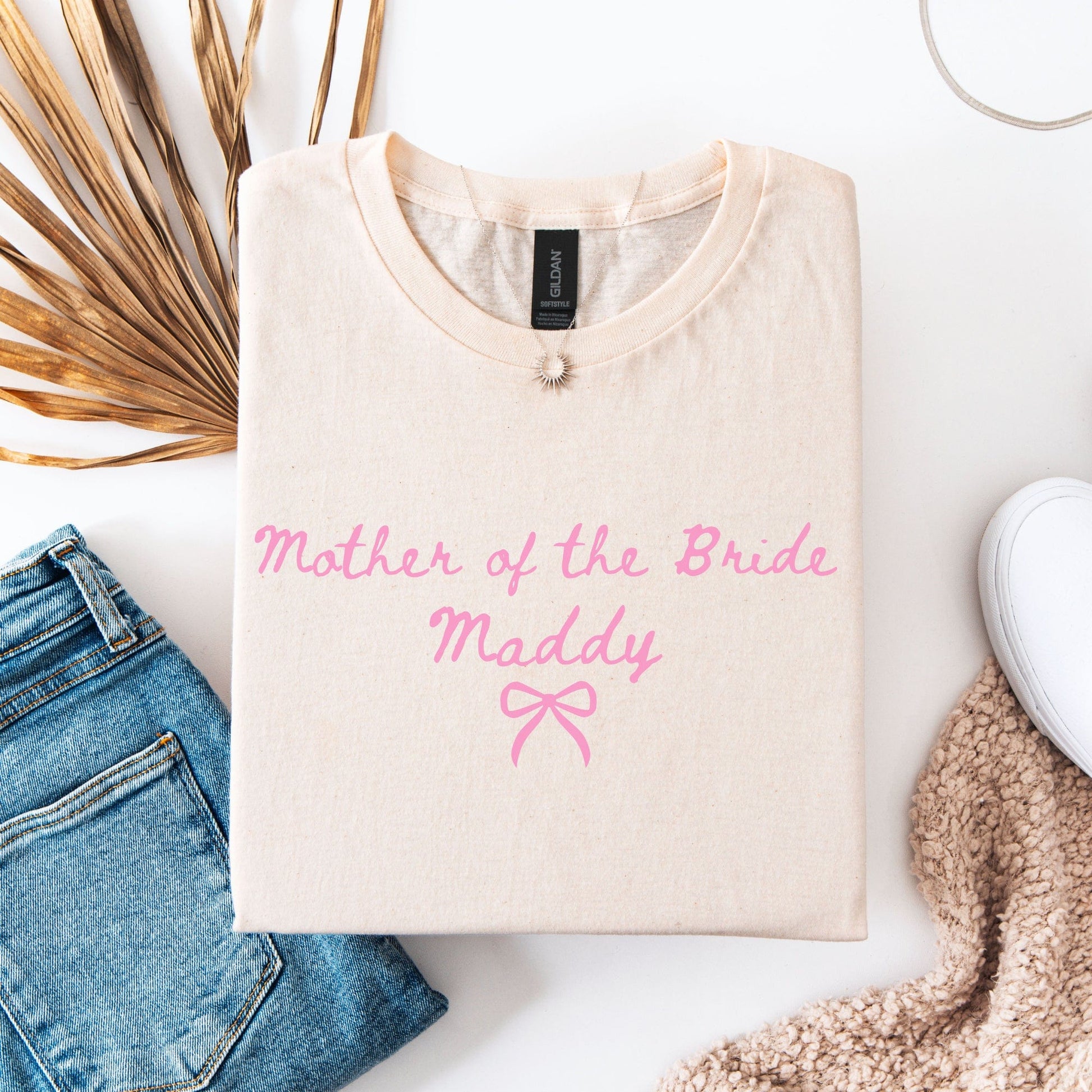 Custom Mother of the Bride Shirt folded cream tee with pink script name Maddy and bow flat lay with jeans and sneakers