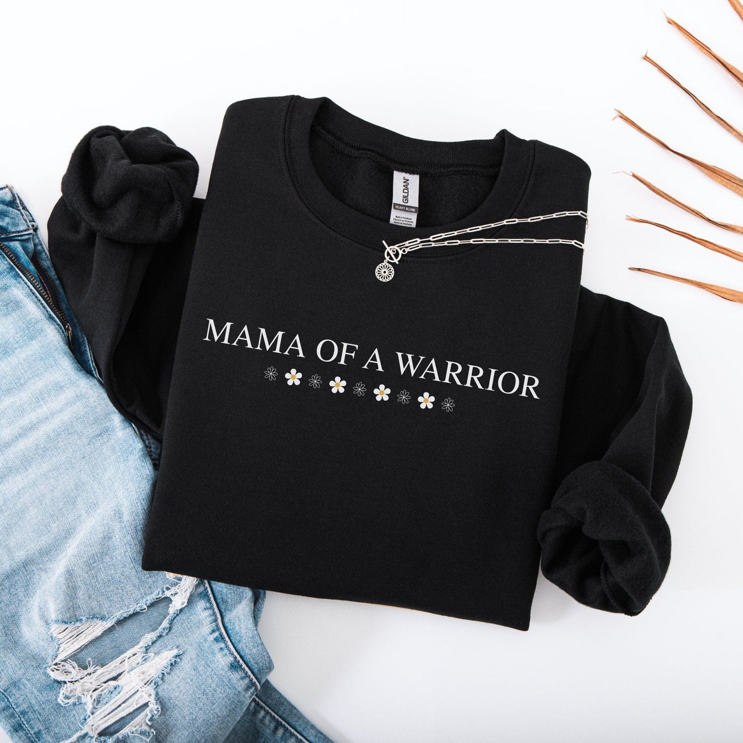 Folded black Mama of a Warrior Sweatshirt with white text and daisy accents styled with ripped jeans and pendant necklaces