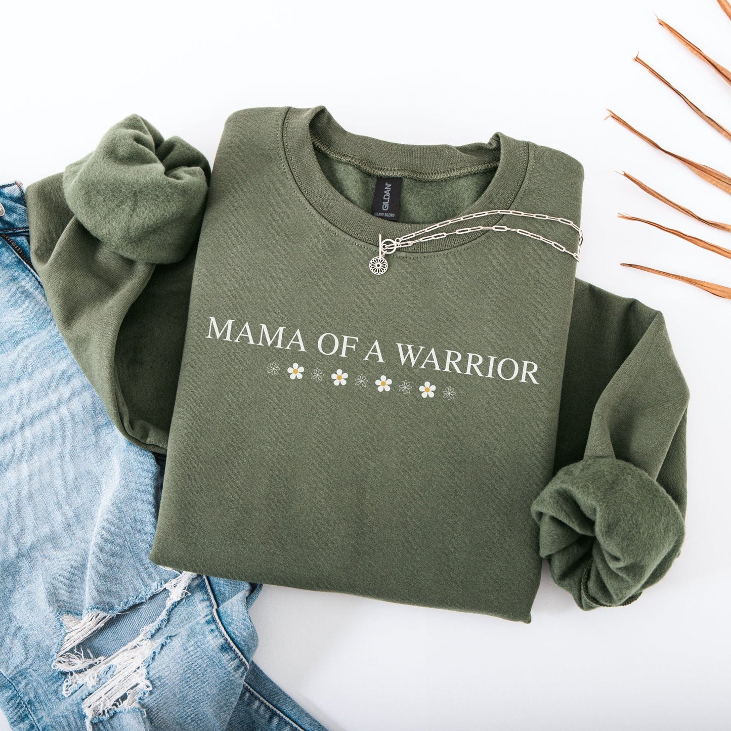 Mama of a Warrior Sweatshirt olive green folded with white floral accents and silver necklace beside ripped blue jeans