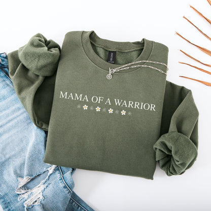 Mama of a Warrior Sweatshirt olive green folded with white floral accents and silver necklace beside ripped blue jeans