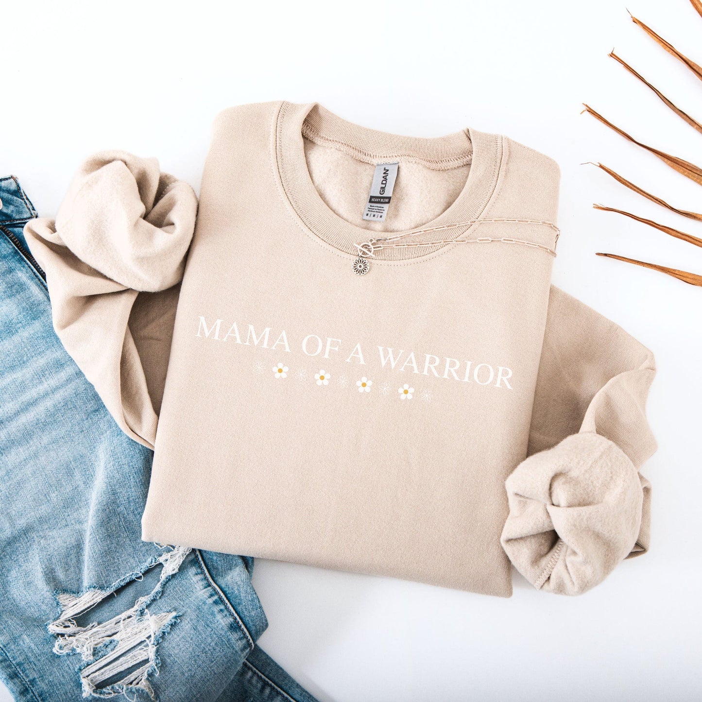 Mama of a Warrior Sweatshirt beige crewneck with white text and daisy design folded with ripped jeans