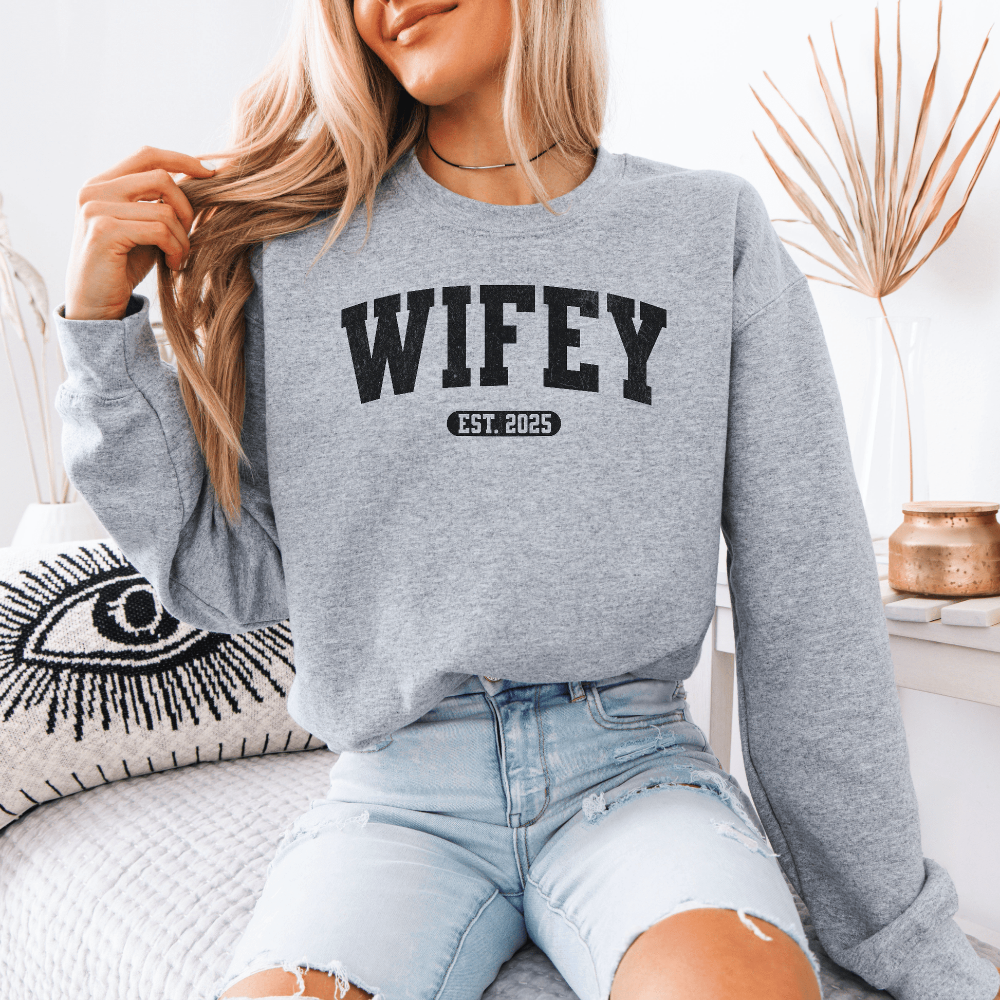 Blonde woman wearing gray custom wifey sweatshirt with WIFEY EST 2025 print sitting on couch in distressed denim shorts