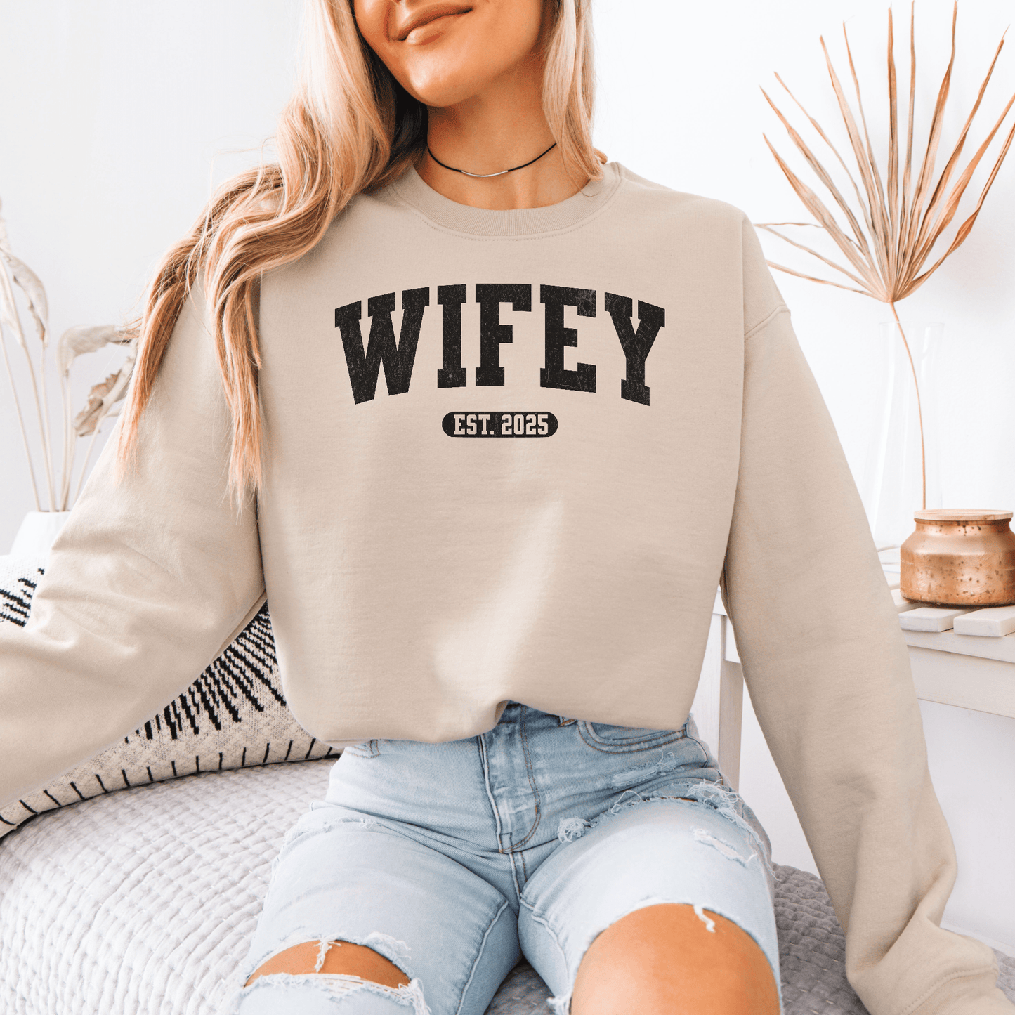 Smiling woman wearing beige custom wifey sweatshirt with WIFEY EST 2025 print and ripped jeans