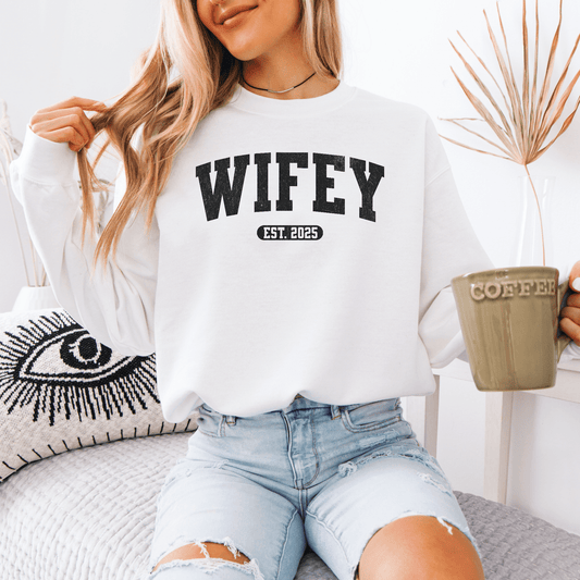 Woman wearing white custom wifey sweatshirt with WIFEY EST 2025 print holding coffee mug on couch