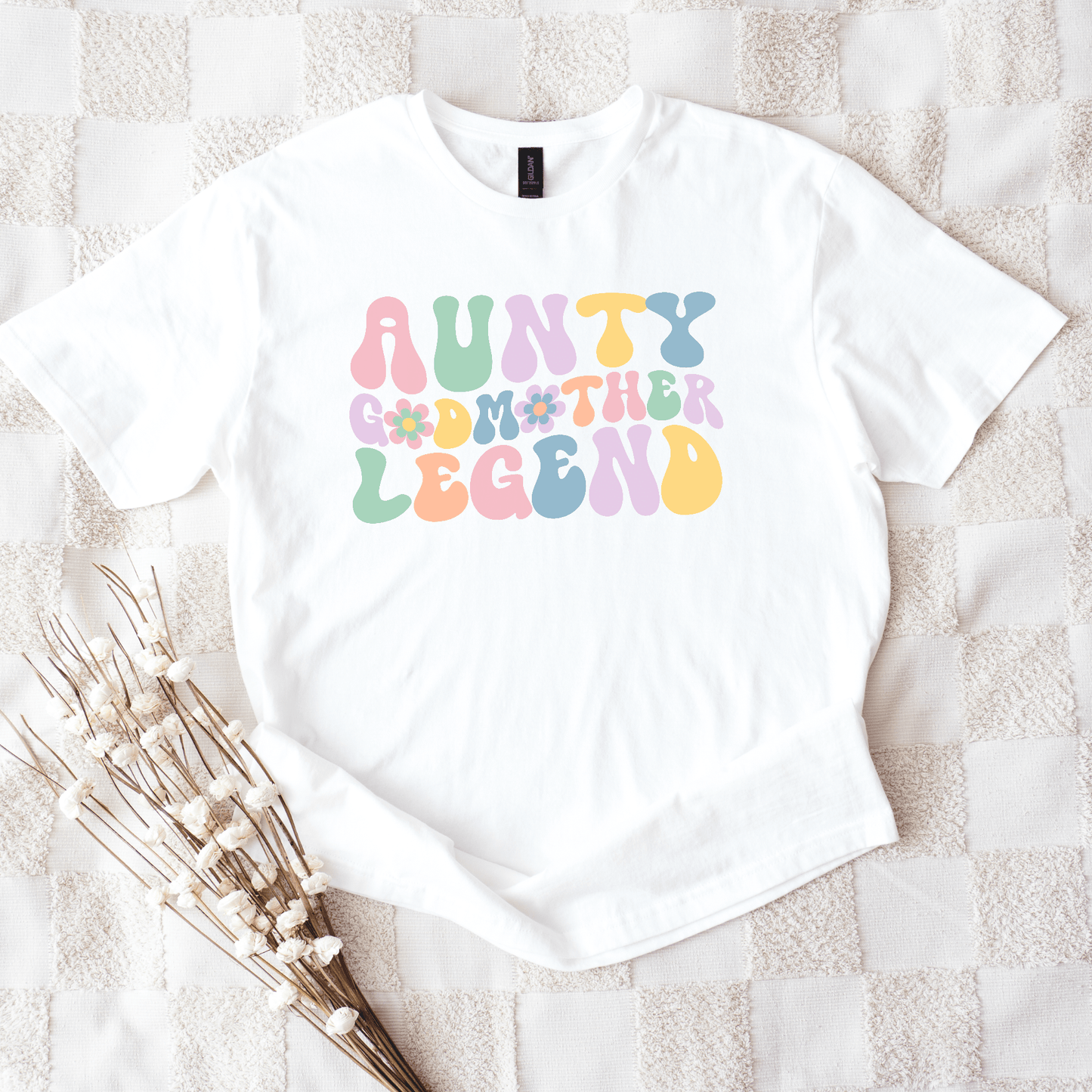 White t shirt with pastel groovy print reading aunty godmother legend shirt laid flat with dried flowers