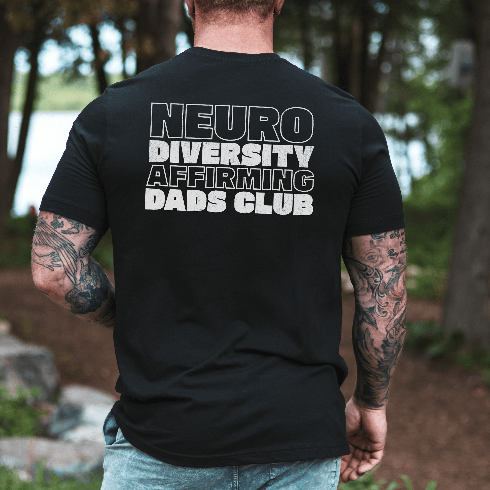 Back view of tattooed dad wearing black Neurodiversity Affirming Dads Club Shirt . bold white lettering outdoor setting