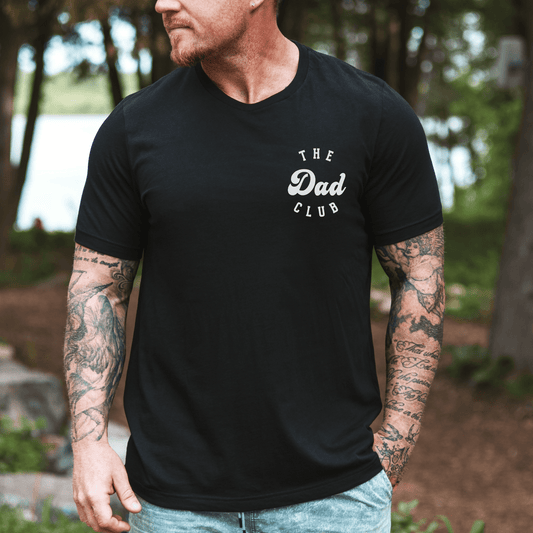 Man wearing black Cool Dads Club Shirt with small chest logo standing outdoors by trees showing tattooed arms
