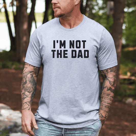 Tattooed man wearing a light gray funny I'm not the dad shirt with bold black text outdoors