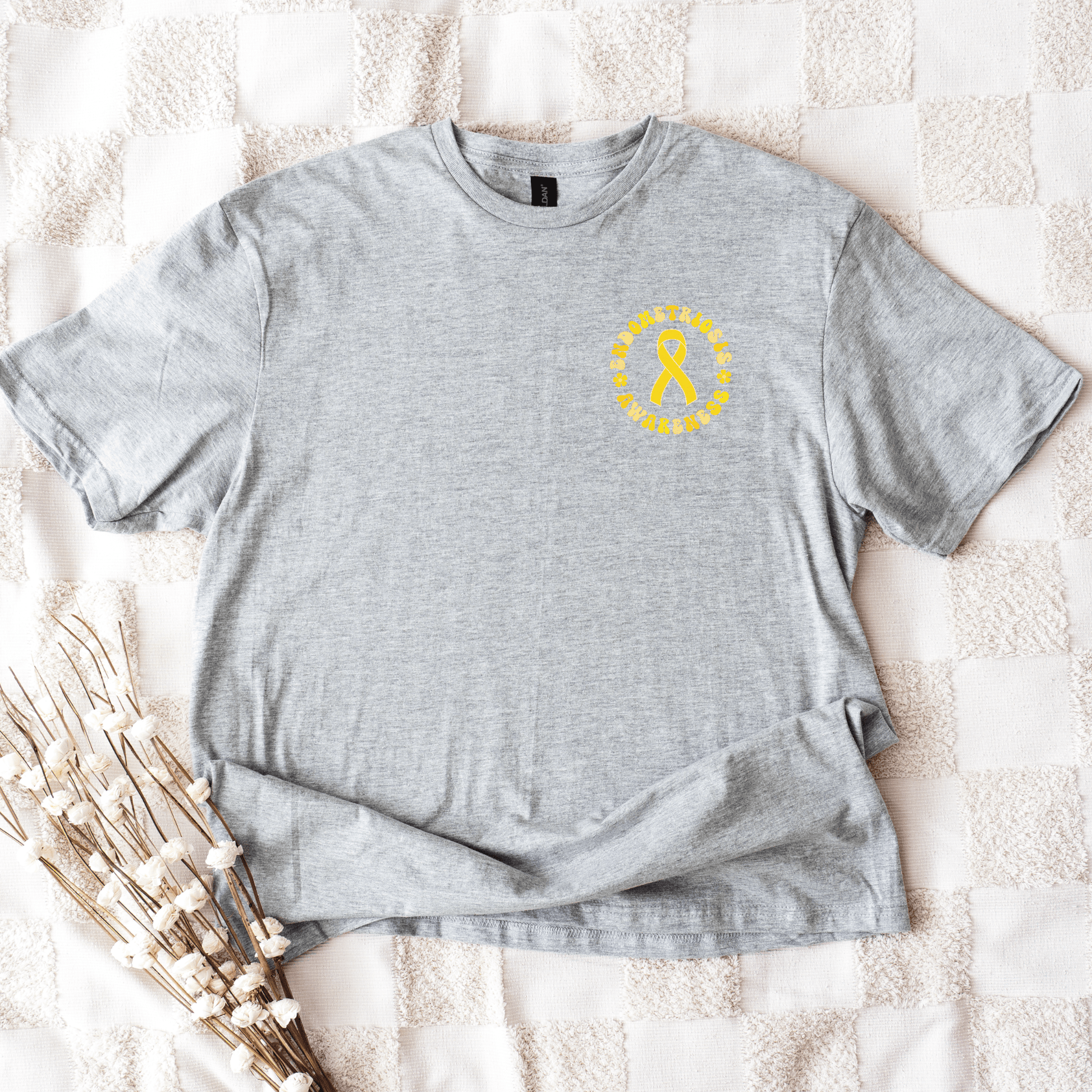 Maddy and George Designs gray tee with small yellow ribbon logo on chest Endometriosis Awareness Shirt on a textured blanket