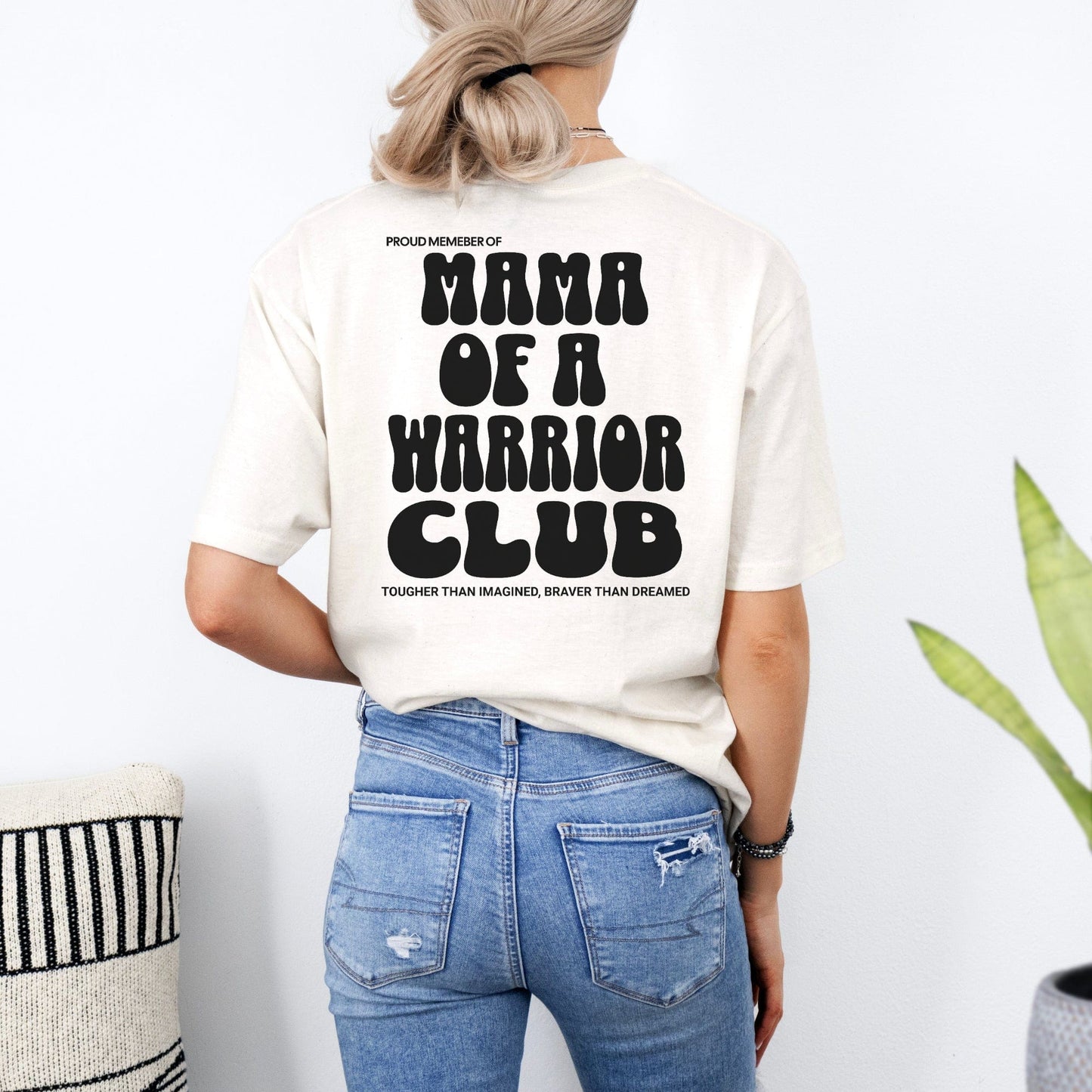 Woman wearing cream tee with black retro print Mama of a Warrior Club Shirt on back paired with blue jeans