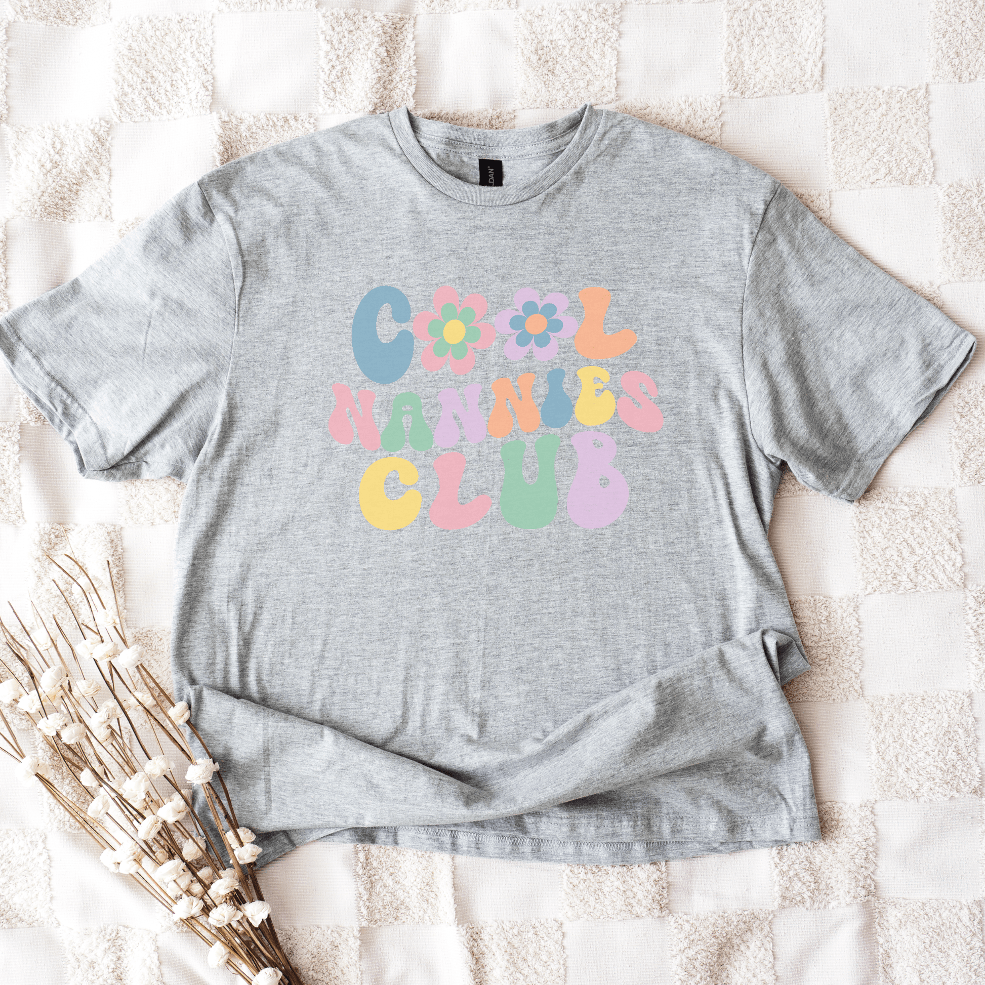 Gray unisex t shirt laid flat on blanket with pastel retro bubble lettering and flower graphics Cool Nannies Club Shirt