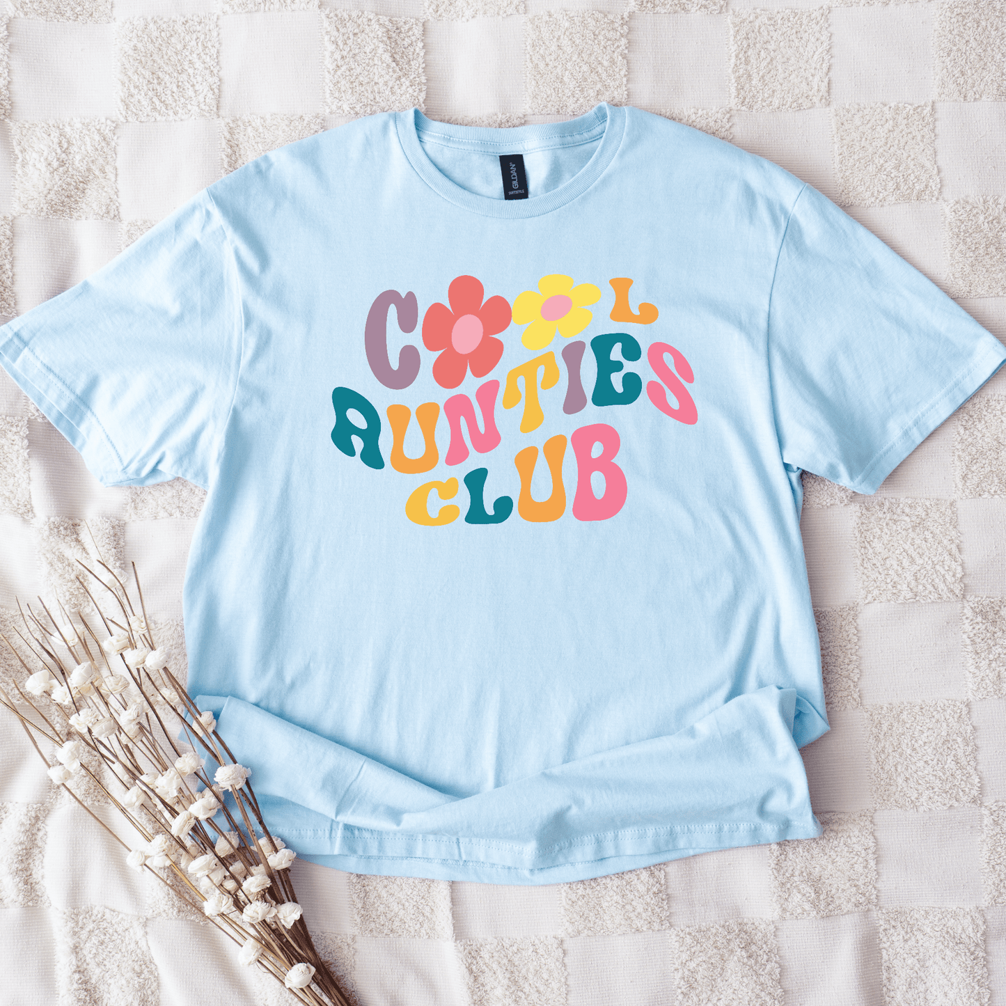Retro Cool Aunties Club Shirt light blue tee laid flat with colorful retro flower lettering on textured blanket