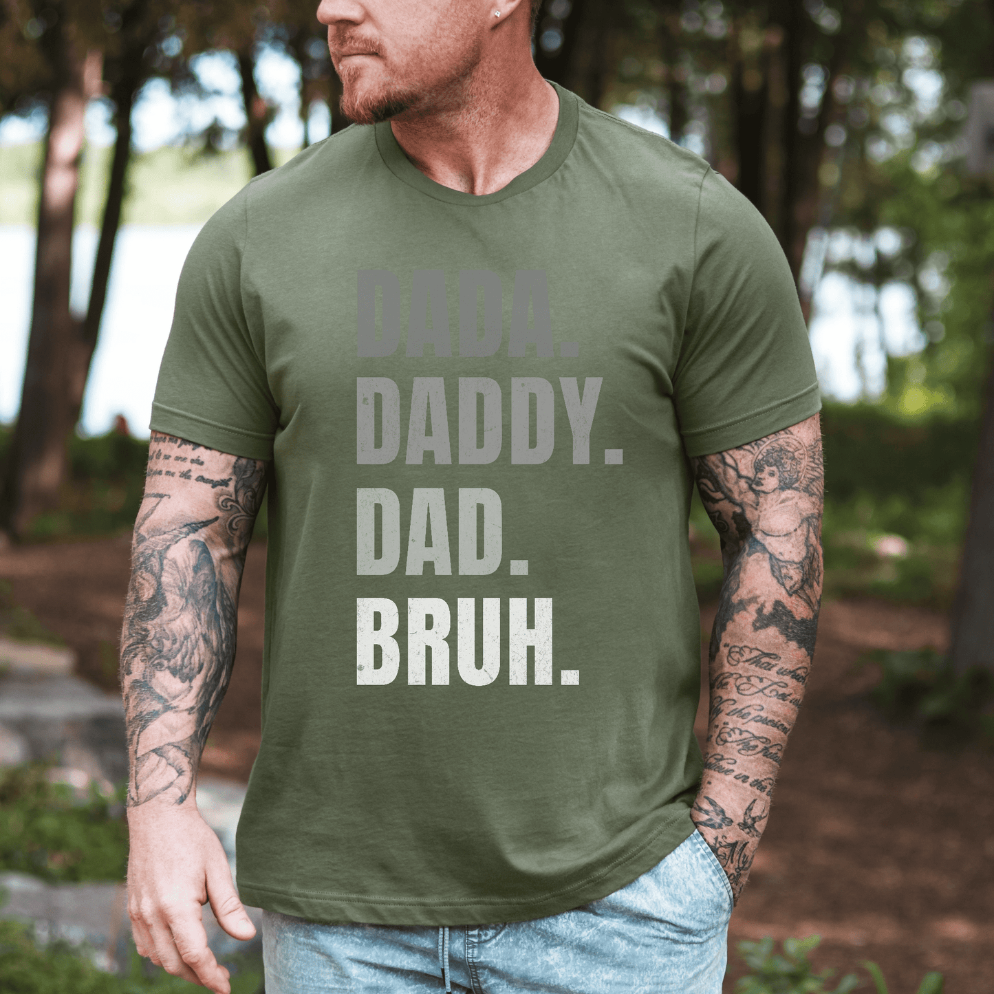 Maddy and George Designs Dada. Daddy. Dad. Bruh Shirt. on tattooed man wearing olive tee outdoors by lake
