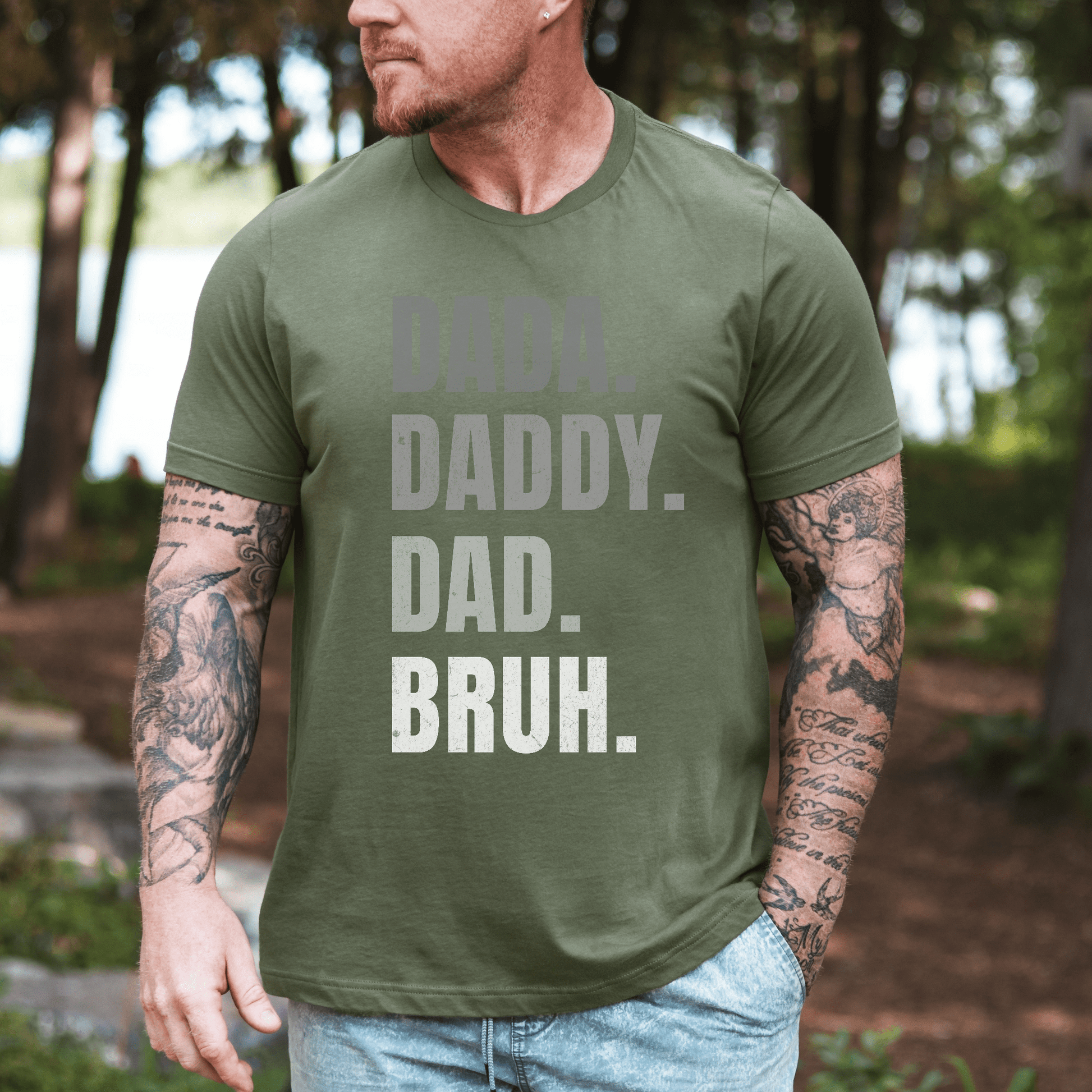 Maddy and George Designs Dada. Daddy. Dad. Bruh Shirt. on tattooed man wearing olive tee outdoors by lake