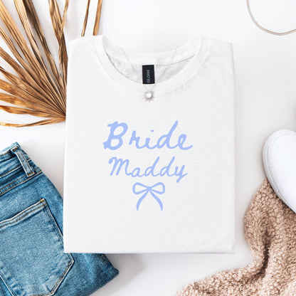 Custom Bride T-Shirt white folded with lavender Bride Maddy script and bow beside jeans and cozy sweater