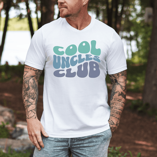 Tattooed man wearing white Cool Uncles Club T-Shirt . retro green and blue bubble letters outdoors by lake