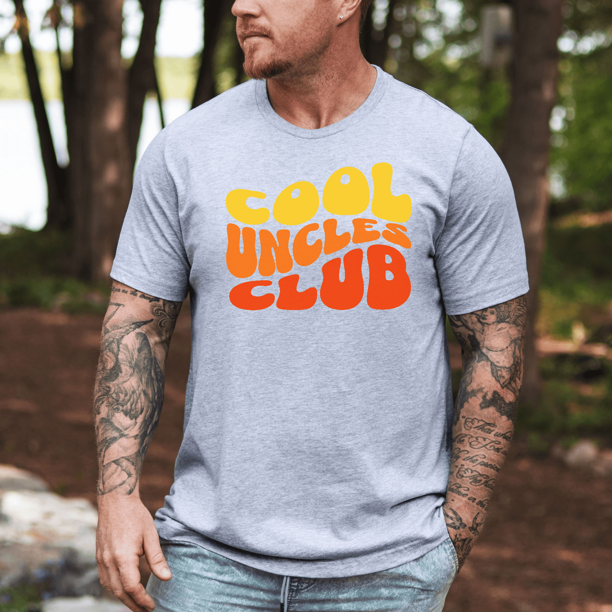 Man wearing gray Cool Uncles Club Shirt with yellow and orange retro bubble text outdoors by woods and lake